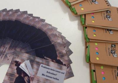 Ein junger Mann sitzt an einem Holztisch und hält ein Tablet in den Händen. Auf dem Bildschirm ist eine bunte Lern-App mit spielerischen Grafiken zu sehen, die Abkürzungen von Flughäfen anzeigen.