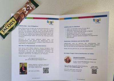 Auf einem Tisch liegt ein aufgeklapptes Faltblatt mit Informationen zum Projekt #ABCforJobs. Daneben liegt ein Cappuccino der Marke Jacobs.
