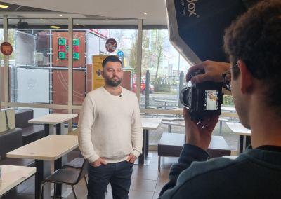 Ein Mann steht in einem Schnellrestaurant und wird von einem Fotografen mit professioneller Kamera und Beleuchtung aufgenommen.