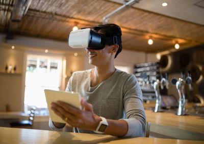 Eine junge Person sitzt in einem Café, trägt eine Virtual-Reality-Brille und hält ein Tablet in den Händen. Sie lächelt und wirkt konzentriert.