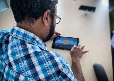 Ein Mann mit kariertem Hemd sitzt an einem Tisch und nutzt ein Tablet. Auf dem Bildschirm ist ein Lernspiel mit einem Flugzeug und Buchstabenkombinationen zu sehen.