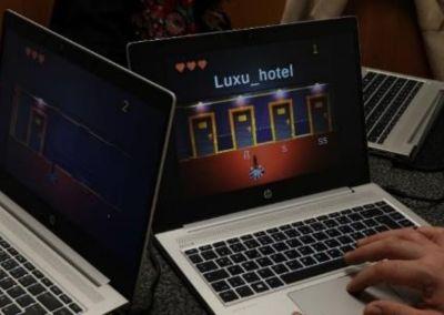 Zwei geöffnete Laptops nebeneinander. Auf einem Bildschirm ist ein Lernspiel mit dem Titel „Luxu_hotel“ zu sehen, das in einer Hotelflurszene spielt.