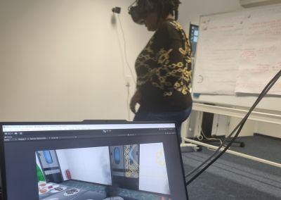 Eine Frau trägt eine VR-Brille und bewegt sich in einem Raum. Auf einem Laptop im Vordergrund ist parallel eine virtuelle Küchenszene sichtbar.