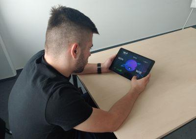 Ein junger Mann sitzt an einem Holztisch und hält ein Tablet in den Händen. Auf dem Bildschirm ist eine bunte Lern-App mit spielerischen Grafiken zu sehen, die Abkürzungen von Flughäfen anzeigen.