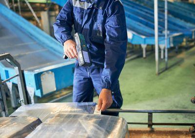 Eine Person in blauer Arbeitskleidung arbeitet in einer Gepäckabfertigungsanlage. Mit einem Handscanner prüft sie Gepäckstücke, die in Folie verpackt sind.