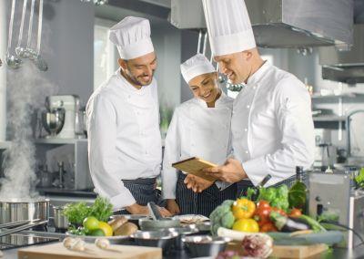 Drei Köchinnen und Köche in weißer Arbeitskleidung stehen in einer Profiküche. Sie lächeln und schauen gemeinsam auf ein Tablet, während frisches Gemüse und Zutaten auf dem Tisch liegen.