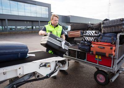Ein Flughafenmitarbeiter in Warnweste lädt Koffer auf ein Transportfahrzeug. Im Hintergrund ist das Flughafengebäude zu sehen.