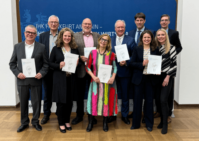 Eine Gruppe von zehn Personen steht in zwei Reihen vor einem blauen Hintergrund mit dem Schriftzug „IHK Frankfurt am Main – Industrie- und Handelskammer“. Alle halten ein Heft mit einem Titelblatt, das eine stilisierte Glühbirne zeigt. In der vorderen Reihe steht eine Frau in einem farbenfrohen, gestreiften Kleid. Die Personen lächeln und präsentieren das Dokument gemeinsam.