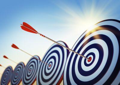 Fünf Zielscheiben mit weißen und blauen Kreisen. In der Mitte steckt jeweils ein roter Pfeil. Im Hintergrund ist blauer Himmel und Sonnenschein zu sehen.