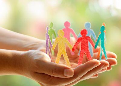 Zwei geöffnete Hände halten mehrere bunte Papierfiguren von Menschen, die sich an den Händen fassen. Die Figuren sind in unterschiedlichen Farben gestaltet und stehen symbolisch für Vielfalt, Zusammenhalt und gegenseitige Unterstützung. Der Hintergrund ist unscharf und in warmen Grüntönen gehalten.