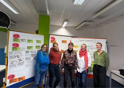 Fünf erwachsene Personen stehen nebeneinander in einem hellen Seminarraum vor zwei großen, mobilen Whiteboards. Auf den Whiteboards sind zahlreiche handschriftliche Notizen, Stichworte und farbige Moderationskarten angebracht. Die Personen wirken locker und zugewandt, einige lächeln in die Kamera. Der Raum ist mit Deckenleuchten ausgestattet, im Hintergrund sind Arbeitsmaterialien und ein grüner Pfeiler zu sehen.