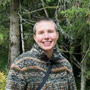 Eine lächelnde Person steht im Freien vor Bäumen.