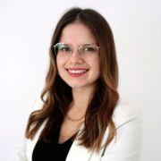 Sally Kiwus aus der Unternehmenskommunikation lächelt mit Brille und weißem Blazer vor hellem Hintergrund in die Kamera.