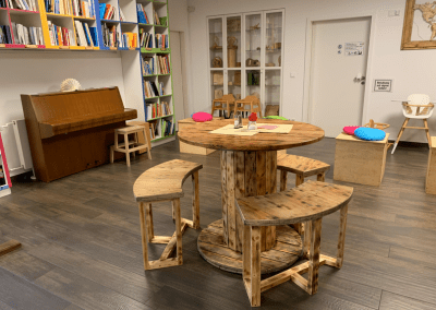 Runder Holztisch mit Sitzbänken im World Café, im Hintergrund Bücherregale und ein Klavier.